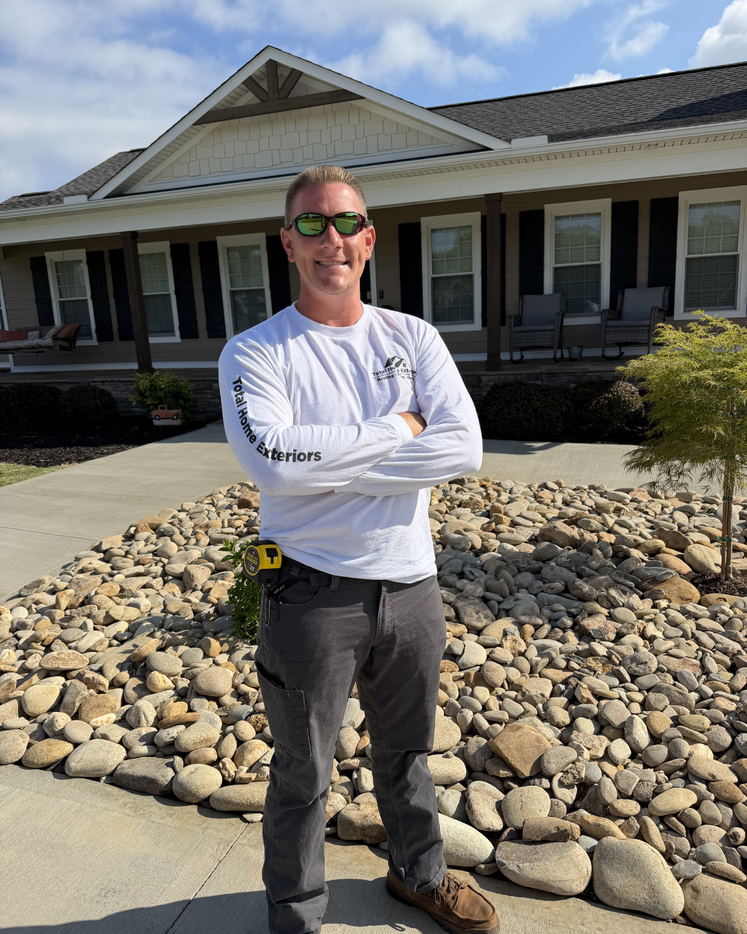 Man standing with arms crossed in front of a house and rock landscaping on a sunny day