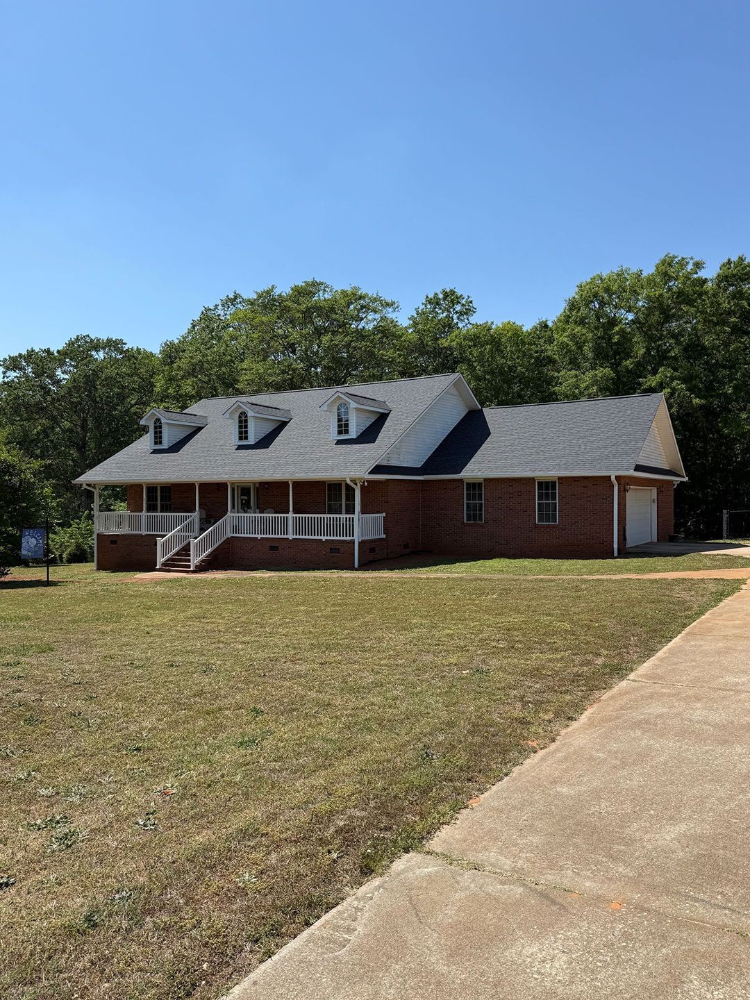 Brick house with gray roof and white porch on a grassy lot beside a sidewalk under a blue sky