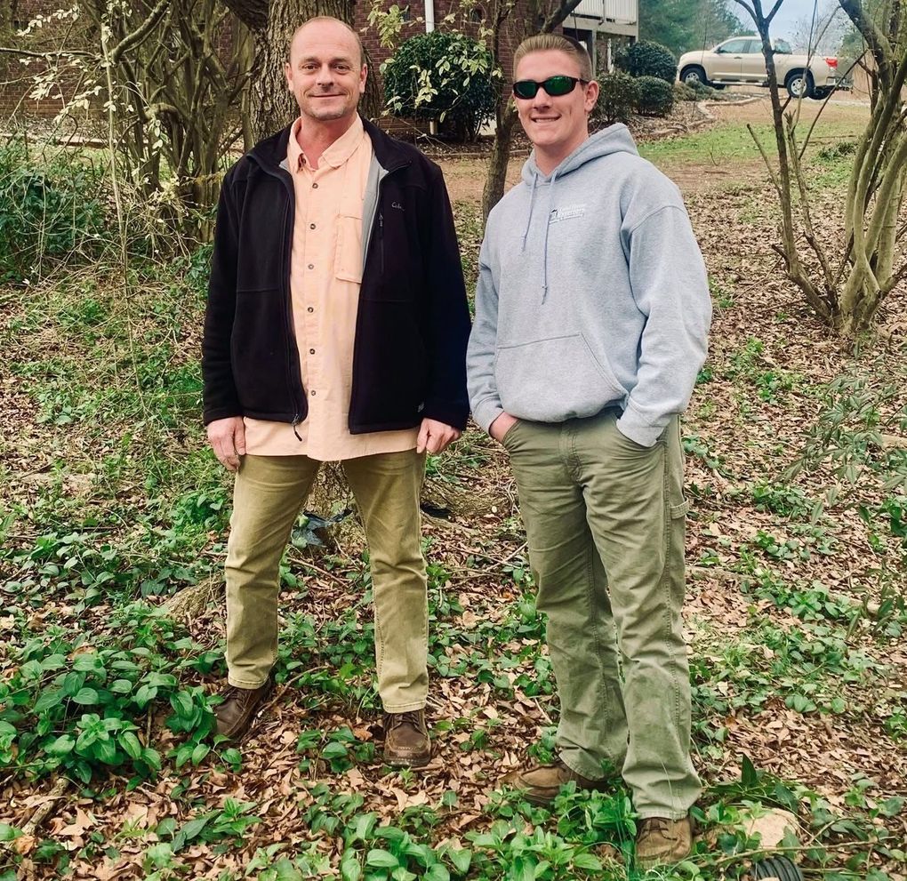 Two men standing in a wooded area; one in a black jacket and the other in a gray hoodie.