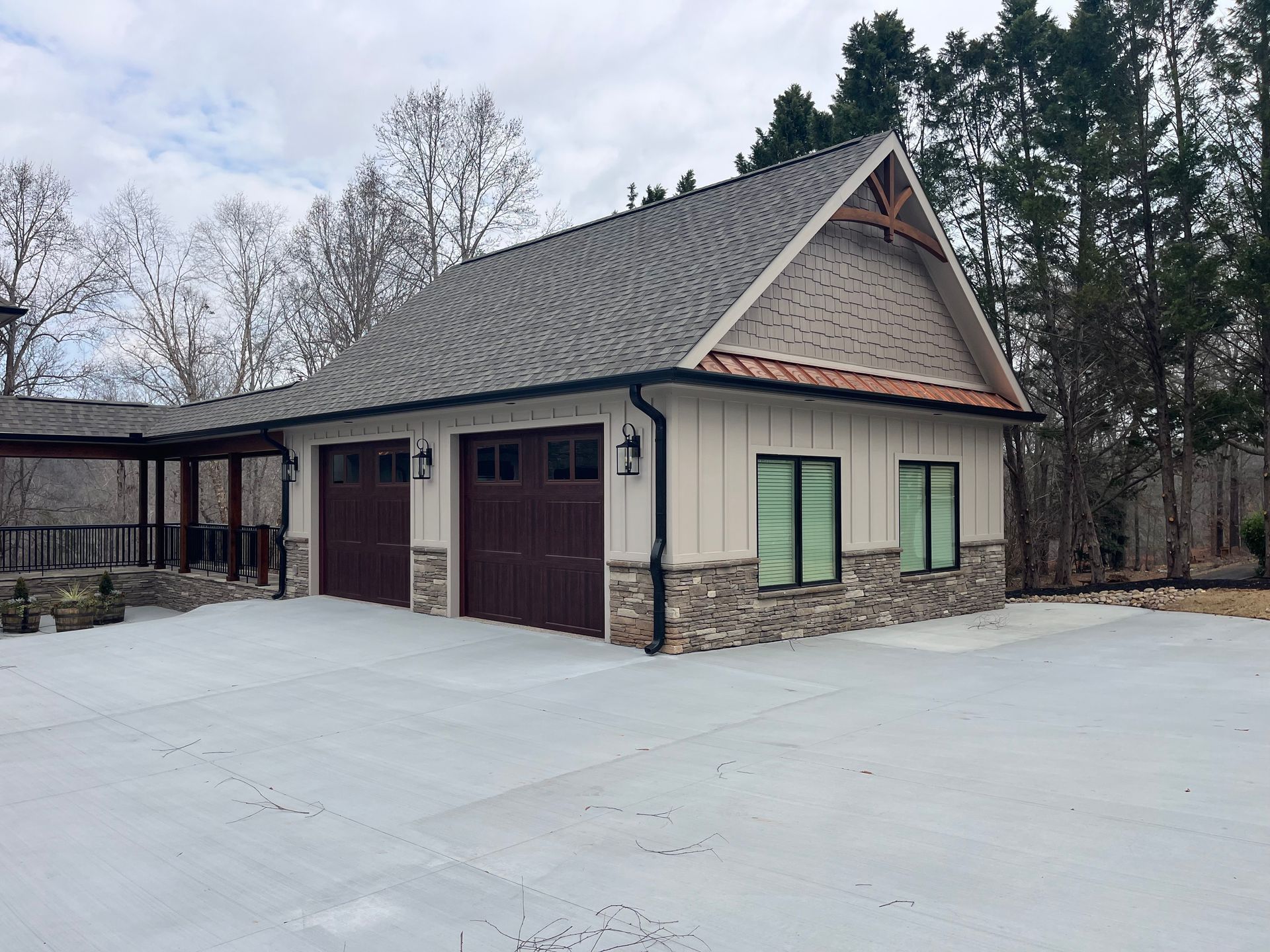 Detached garage with brown doors, green shutters, and a steep roof on a snowy driveway near trees