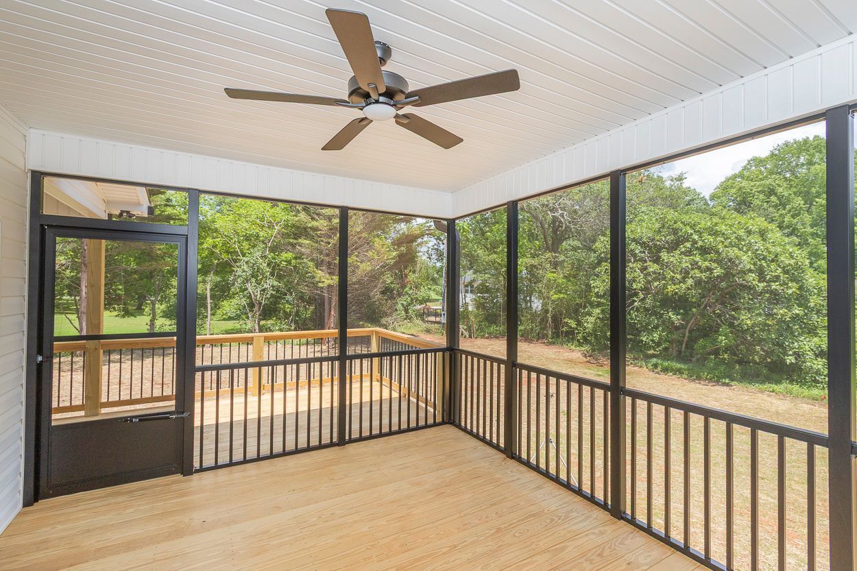Screened-in porch with black frames, wooden railings, and a ceiling fan, overlooking a green outdoor view.