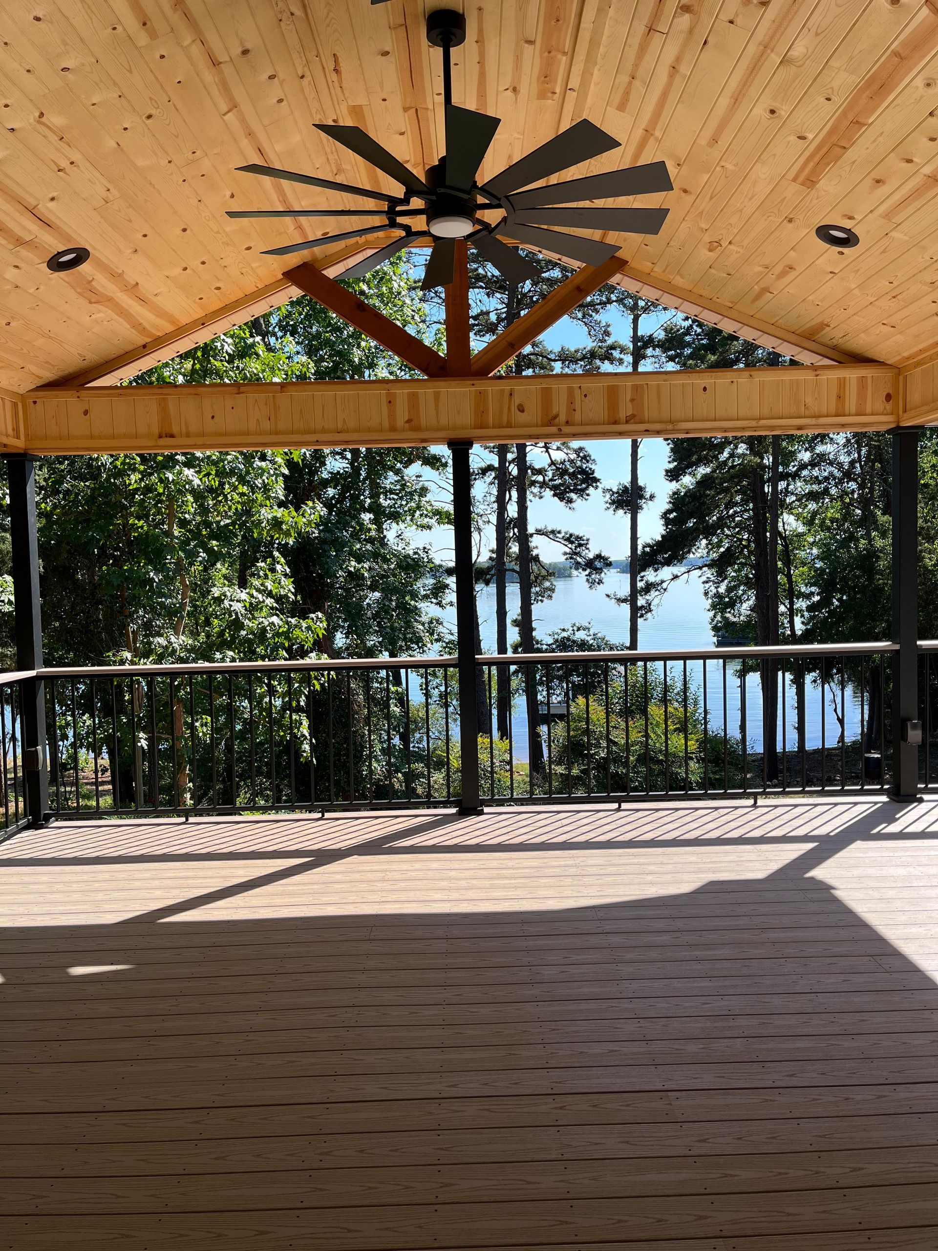 Covered patio with wooden ceiling fan, overlooking trees and a distant lake through black railing.