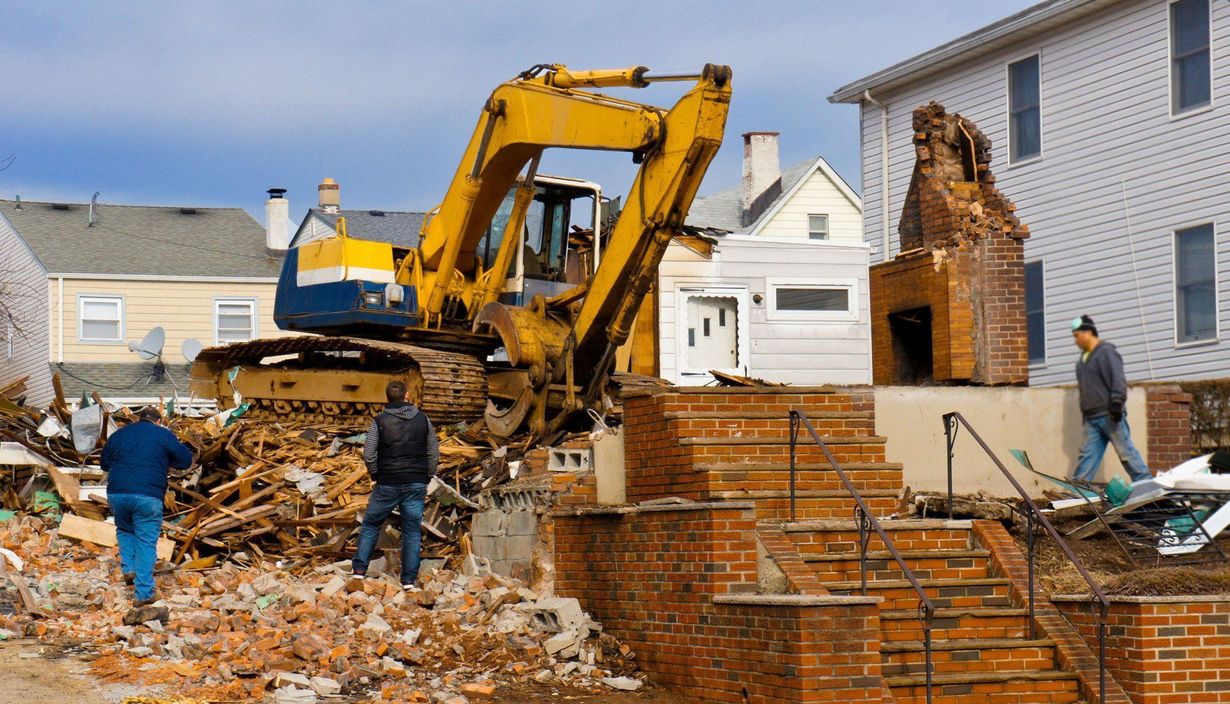 Yellow excavator demolishing a brick house, workers nearby.