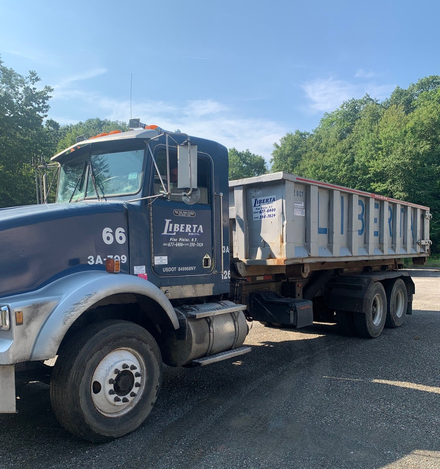 Blue dump truck with trailer parked outside; trees and blue sky background.