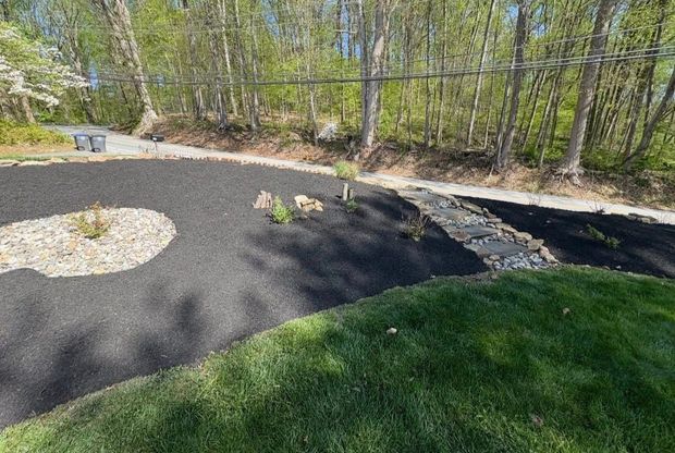Curved landscaped garden bed with black mulch, stepping stones, and green grass beside a wooded path