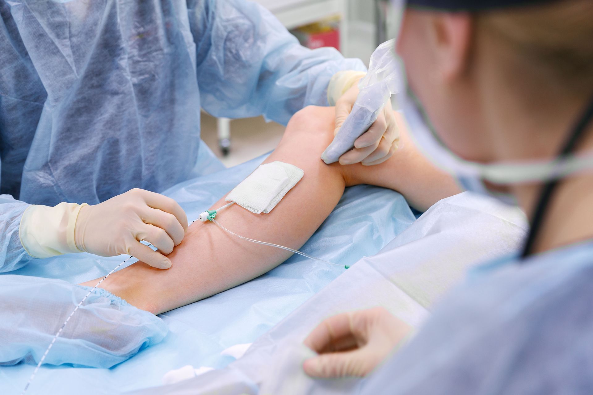Medical professionals performing a procedure on a patient's arm; setting is an operating room.