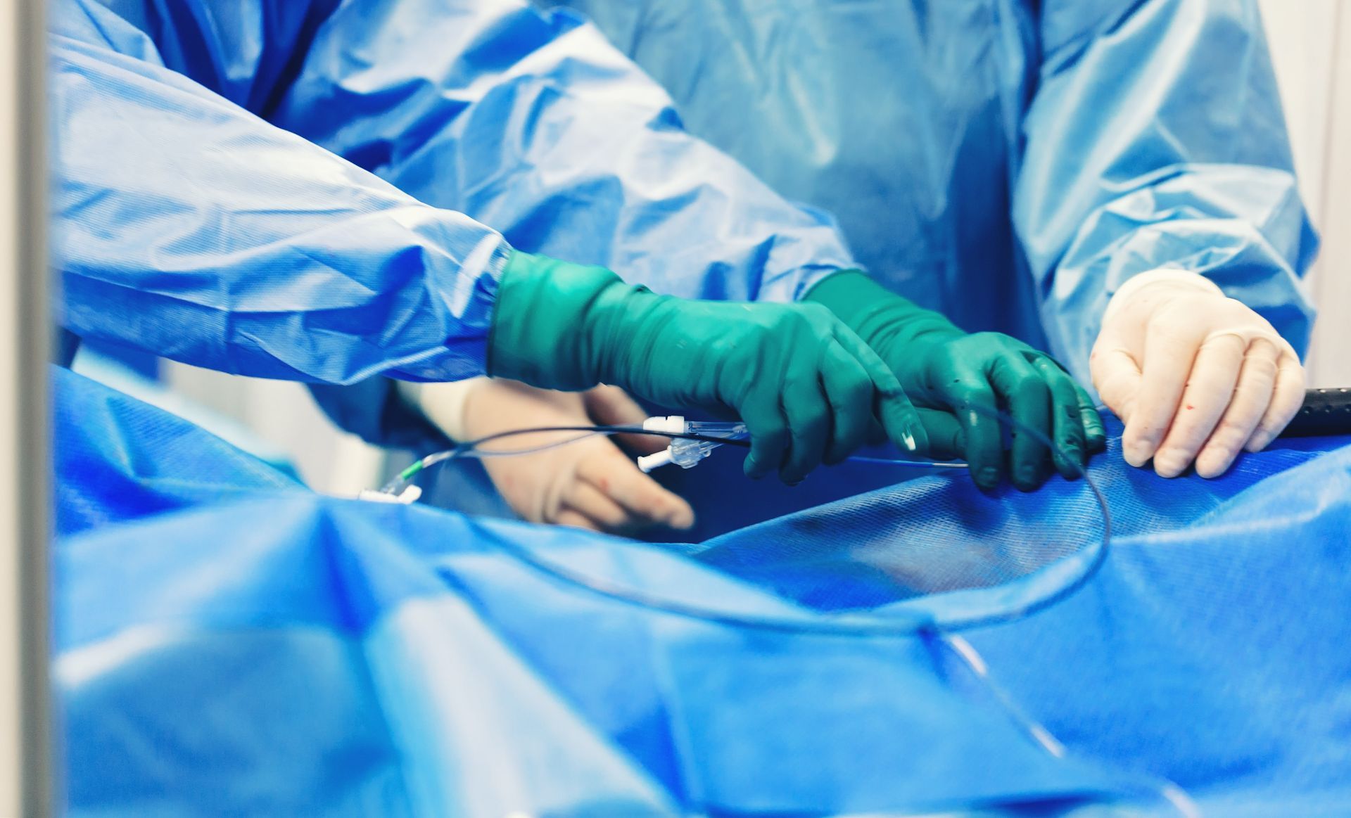 Surgeons in blue scrubs and green gloves operating, hands over patient draped in blue.