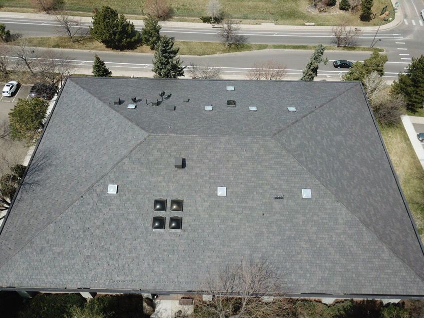 Aerial view of a building with a gray shingle roof, skylights, and vents. Road and trees are in the background.