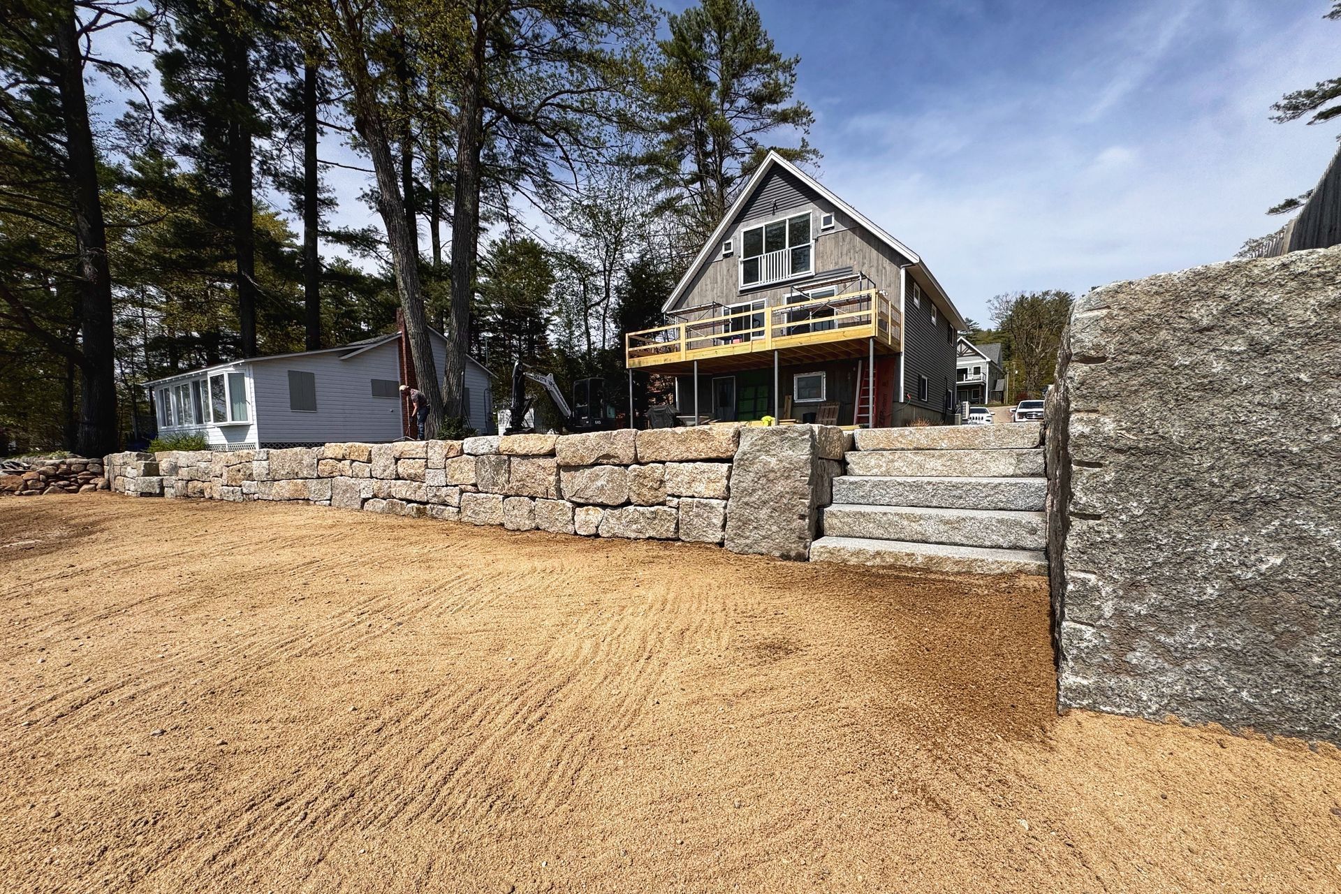 A cottage with a deck and stone wall. Brown gravel ground, and trees in the background.