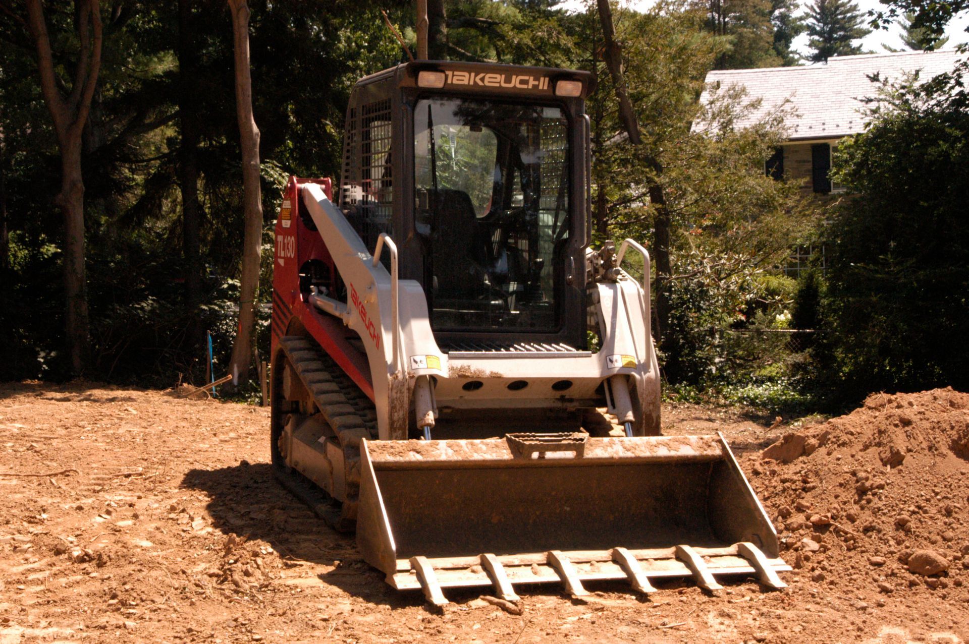 A white and orange track loader with a bucket sits on a dirt plot with a house and trees in the background.