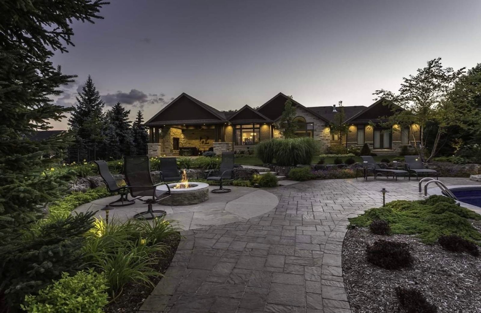 Backyard patio at dusk with fire pit, seating, landscaped gardens, and a house in the background.