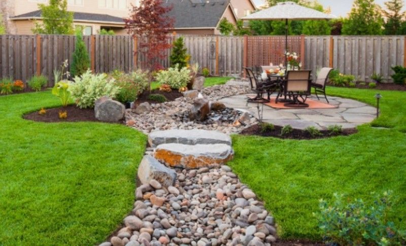Backyard with a rock-lined stream flowing toward a stone patio with a table and chairs, surrounded by greenery and a wooden fence.