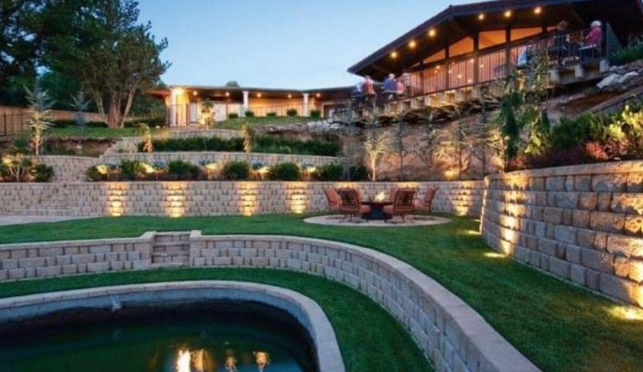 Night view of a terraced outdoor space with illuminated retaining walls, lawn, and water feature near a house with lights.