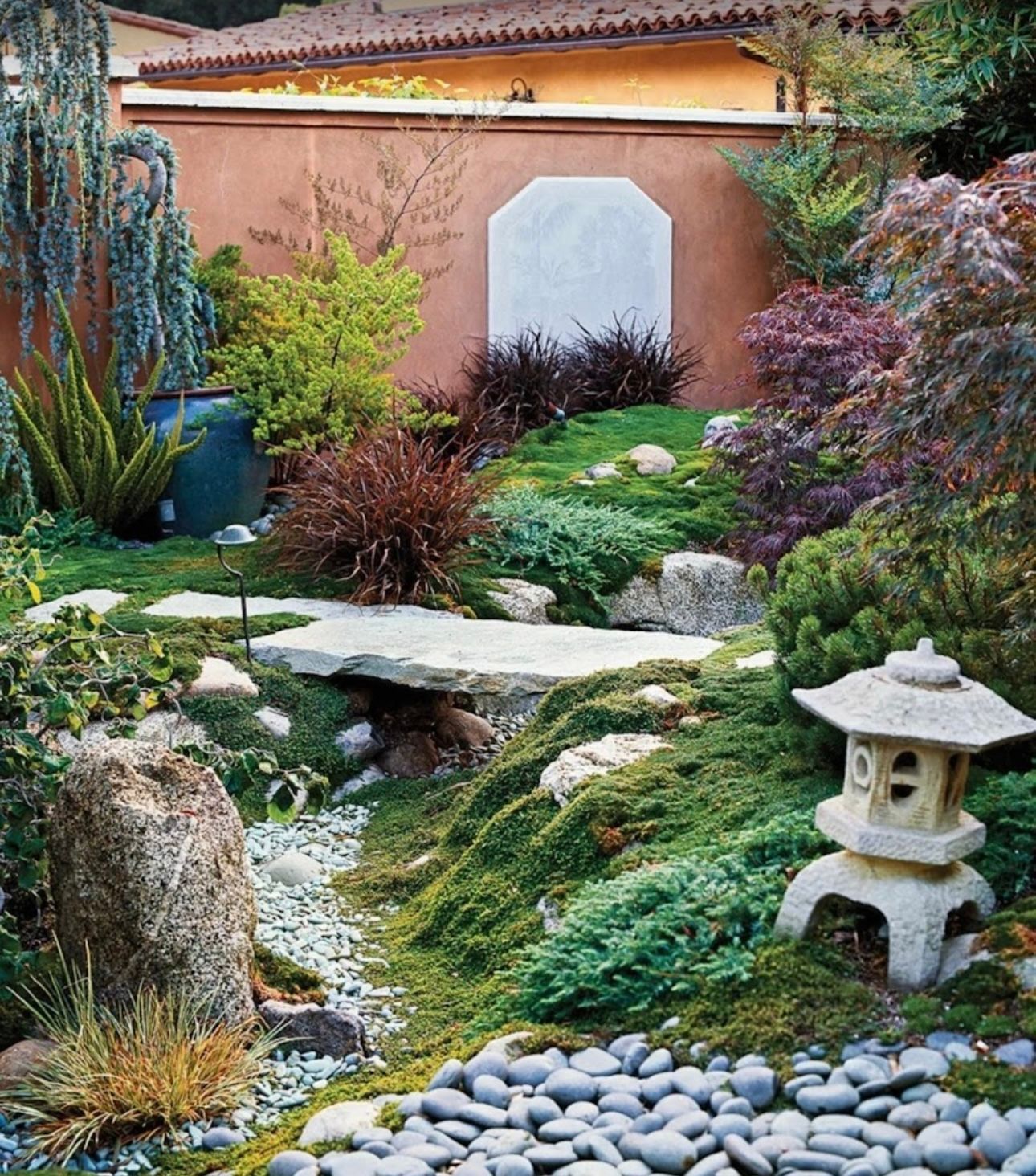 Japanese garden with a stone bridge, lantern, and plants, against a terracotta wall.