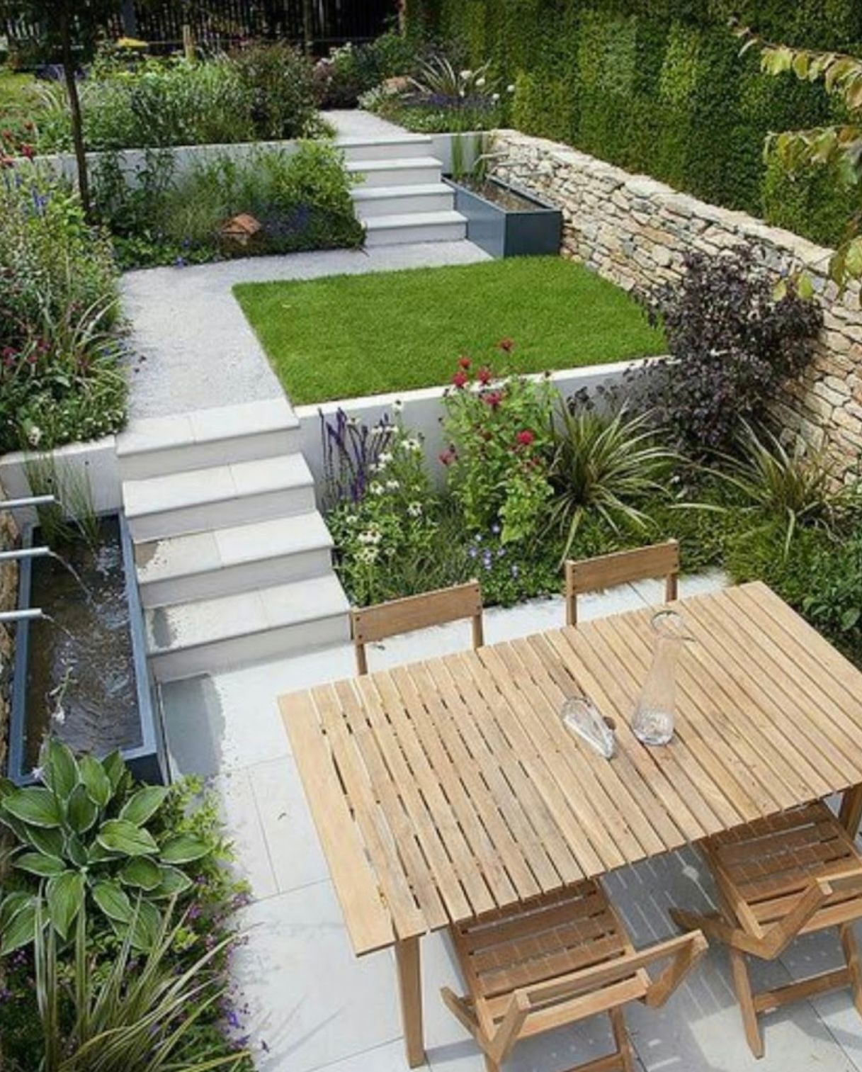 Elevated backyard garden with stone walls, stairs, dining table, and lawn area. Lush greenery and water feature.