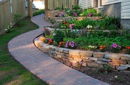 Curving brick pathway through tiered garden beds with stone walls, blooming flowers, and lush greenery.
