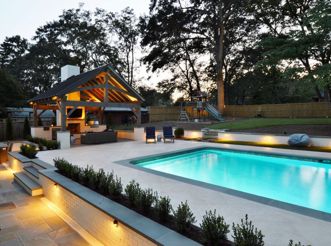 Backyard pool with outdoor kitchen under a wooden pergola, evening scene, lit by soft lights.