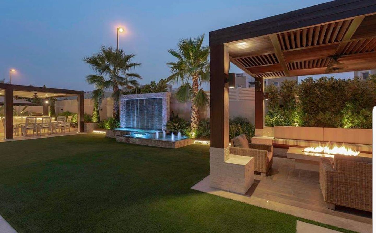 Outdoor evening scene: manicured lawn, water feature, lit pergolas, and fire pit with seating.