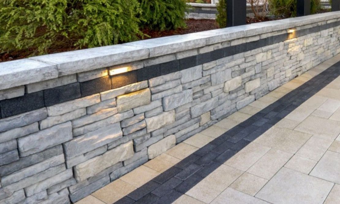 Stone retaining wall with built-in lighting and a decorative dark band, adjacent to a paved patio.