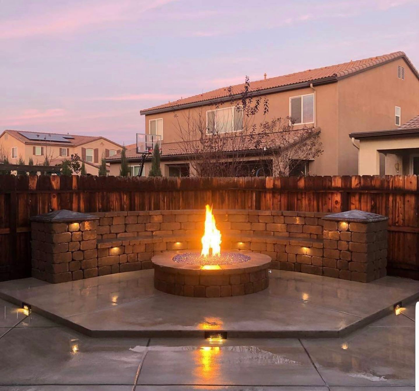 Fire pit in a backyard with a stone wall and built-in lights, under a twilight sky.