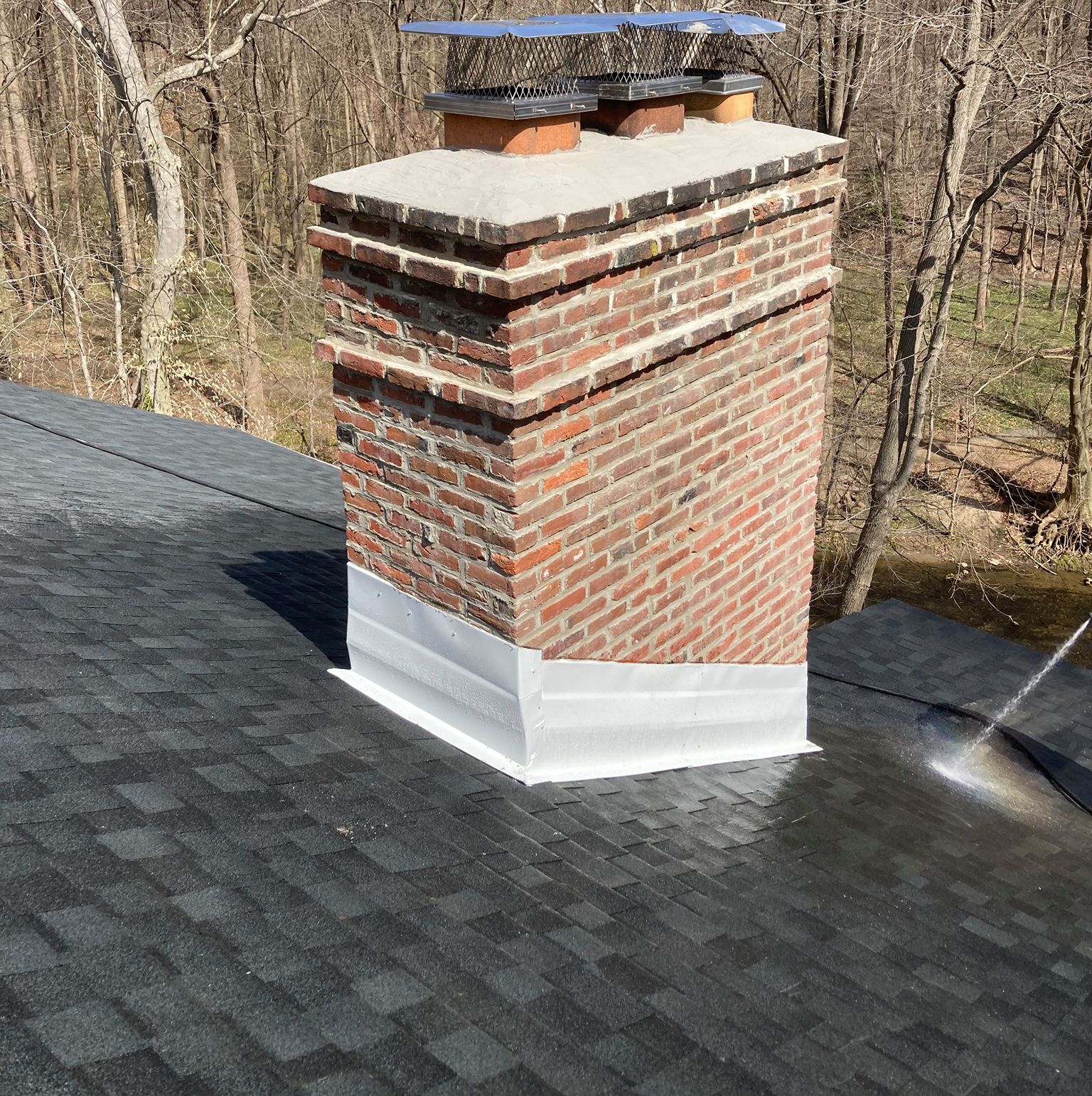 Brick chimney on a dark shingle roof with flashing, set against a wooded backdrop.