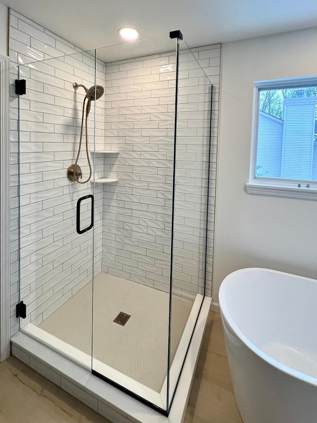 Modern white-tiled bathroom with glass shower, showerhead, handheld sprayer, and bathtub next to a window