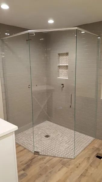 A corner neo-angle glass shower enclosure with gray tiled walls, a built-in shelf, and a patterned tiled floor.