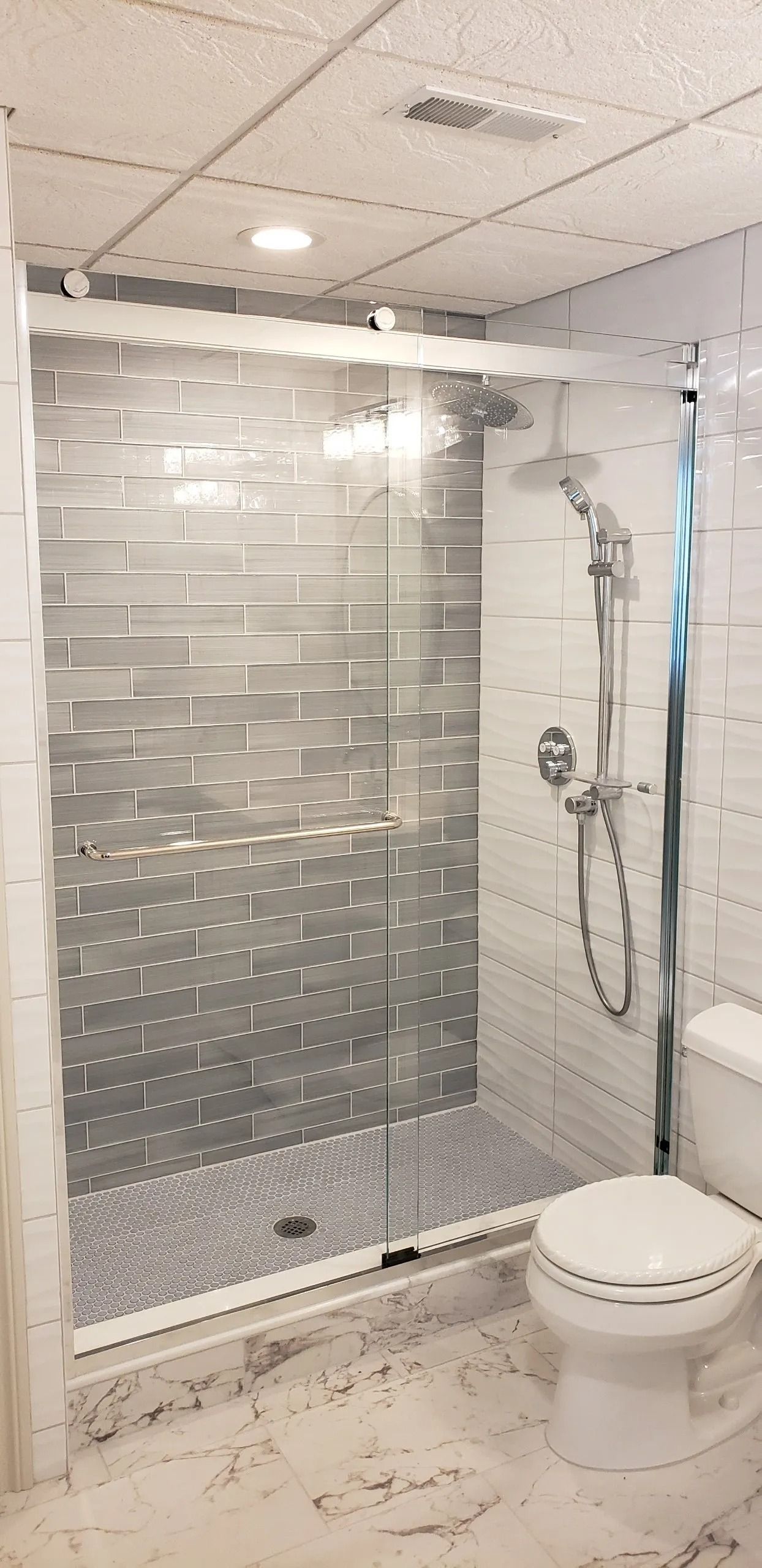 A modern bathroom featuring a walk-in shower with gray-toned tiled walls, a pebble-textured floor, and a glass sliding door.