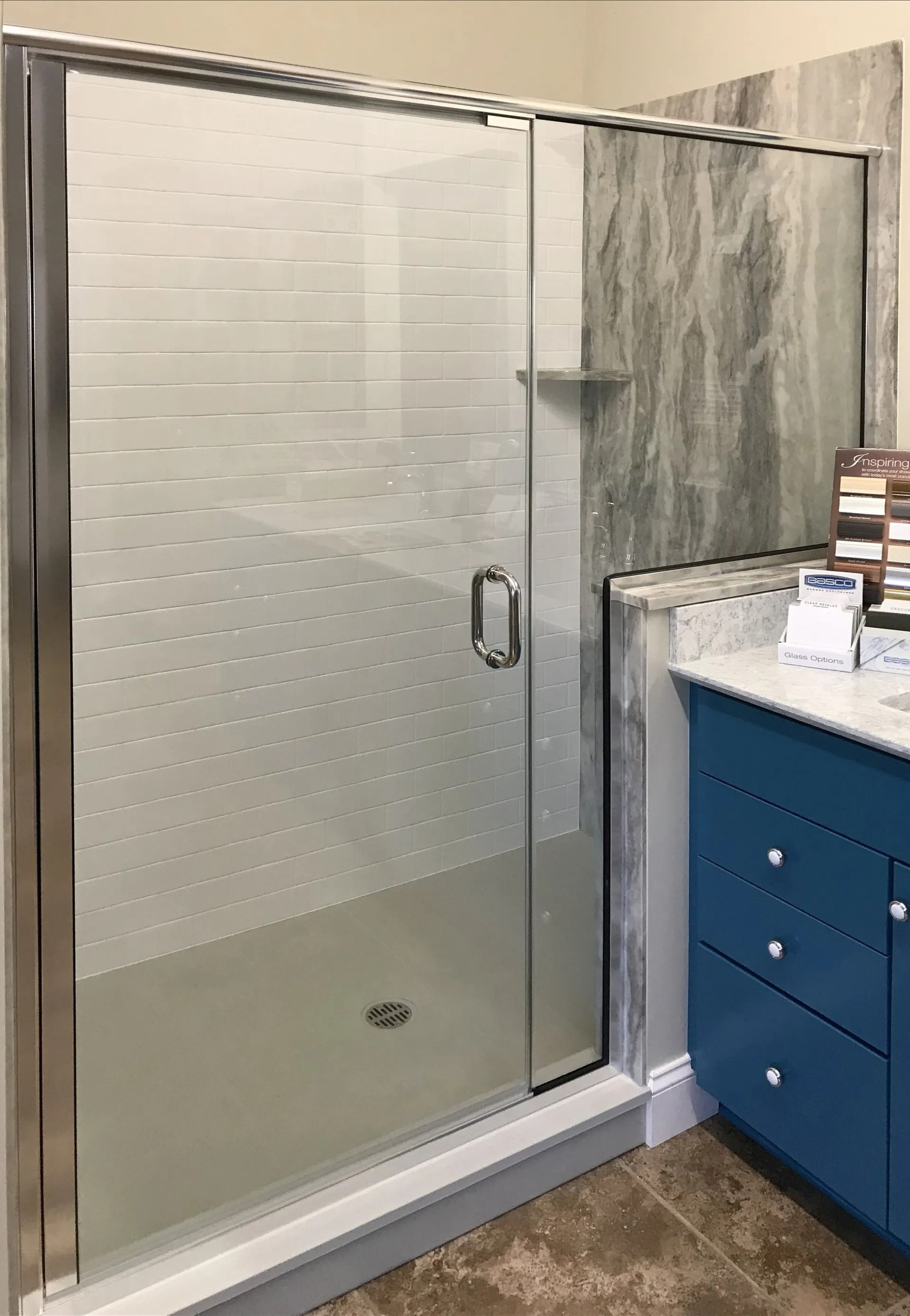 A glass shower door with a striped privacy pattern stands next to a blue bathroom vanity with drawers and a stone top.