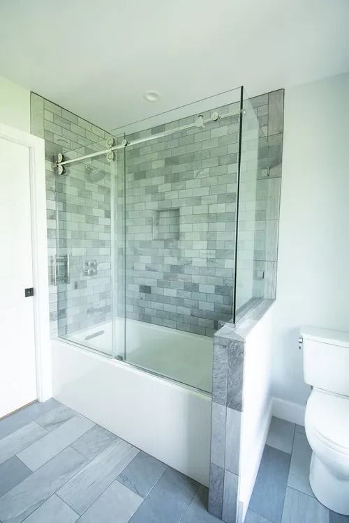 A bathroom featuring a bathtub with a glass shower door, light gray brick-style tile walls, and gray floor tiles.