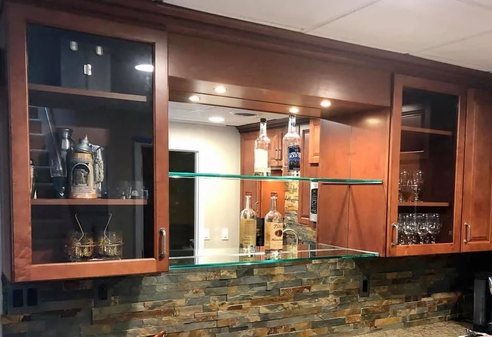 A wooden home bar with glass-front cabinets, glass shelving, and a decorative stone backsplash under recessed lighting.