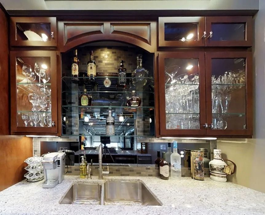 A home bar featuring dark wood cabinets, a mirrored backsplash, glass shelves, a double sink, and various liquor bottles.