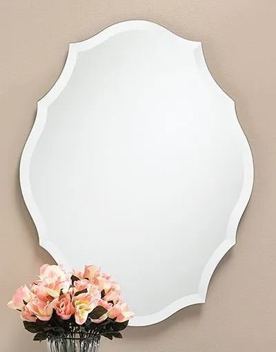 A wall-mounted mirror with a scalloped, frameless edge hanging above a vase of soft pink artificial roses.