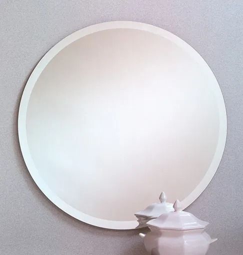 A large, circular, frameless mirror mounted on a light gray wall, with a white decorative lidded bowl in the foreground.