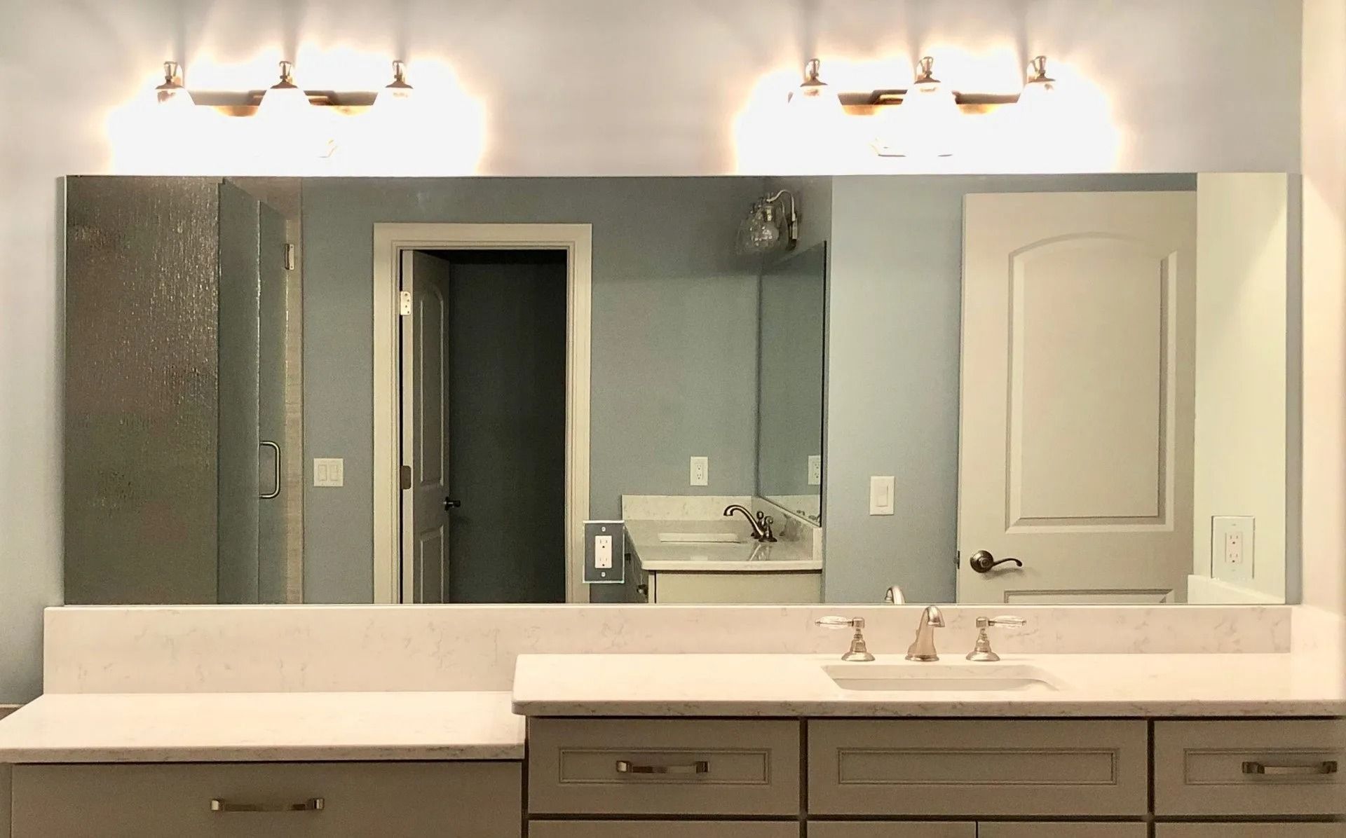 A bathroom vanity with a long rectangular mirror, two wall-mounted light fixtures, white countertops, and gray cabinets.