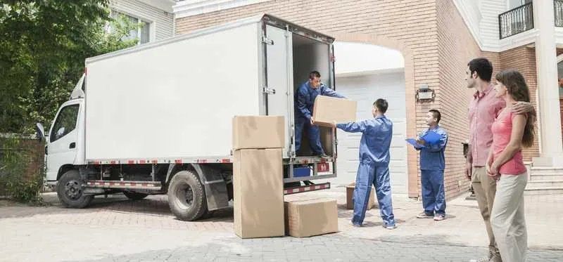 Movers unloading boxes from a truck in front of a house while a couple watches.