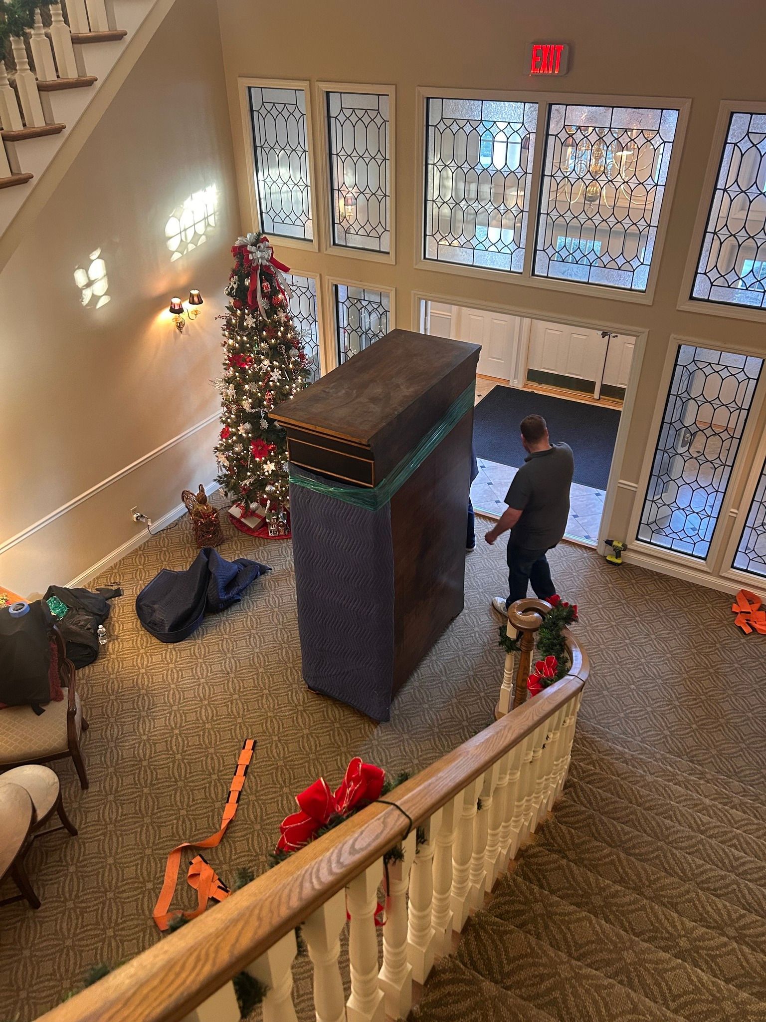 A man moving a large, wrapped object inside a building with a Christmas tree and staircase.