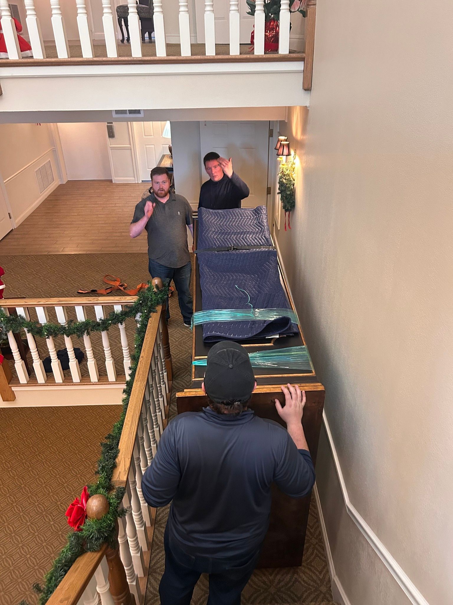 Three men moving a large, decorated object down a staircase; Christmas garland visible.