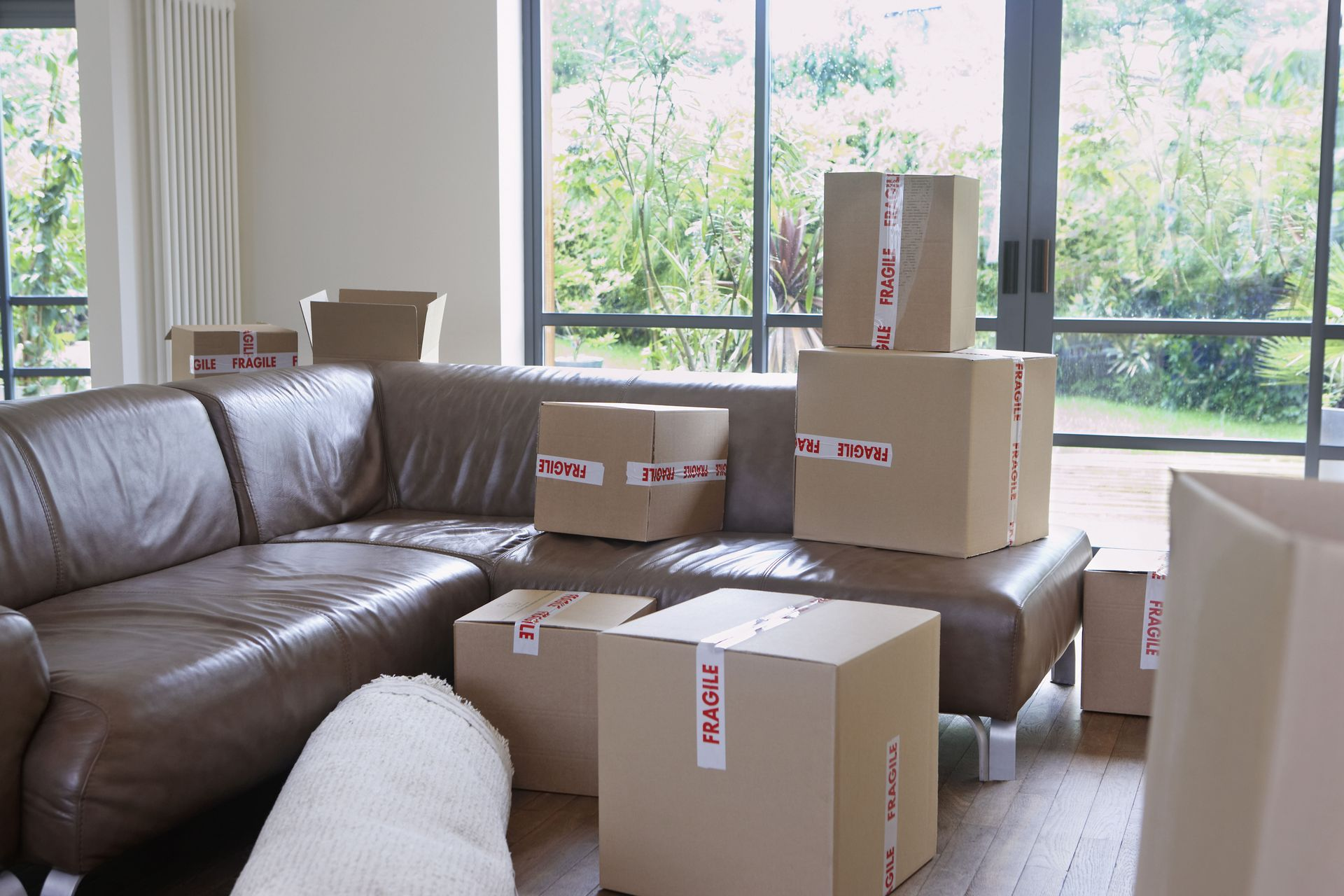 Cardboard boxes on a brown leather sectional couch near a large window, indicating a move.