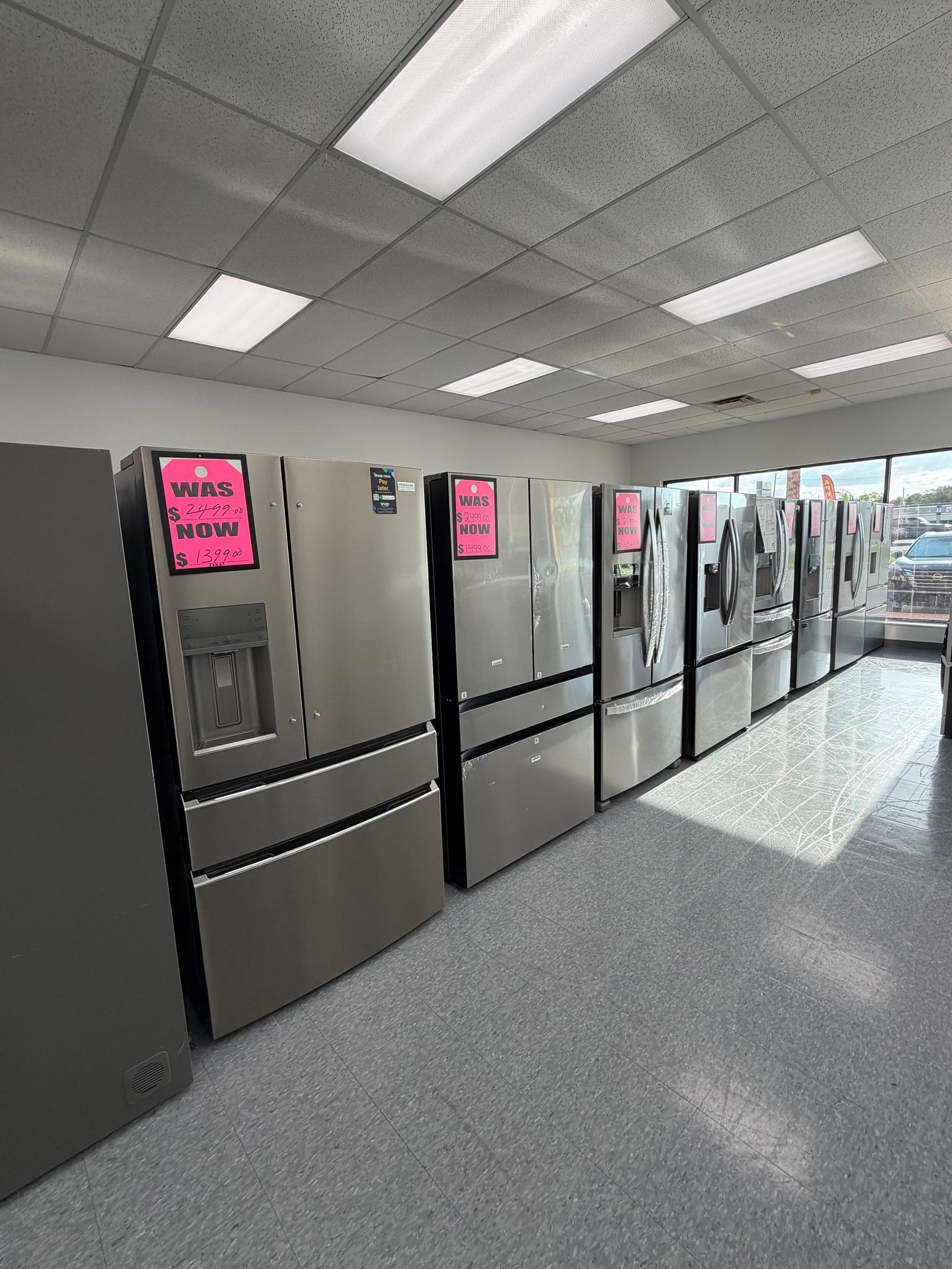 Row of stainless steel refrigerators in a bright store aisle with pink sale signs.