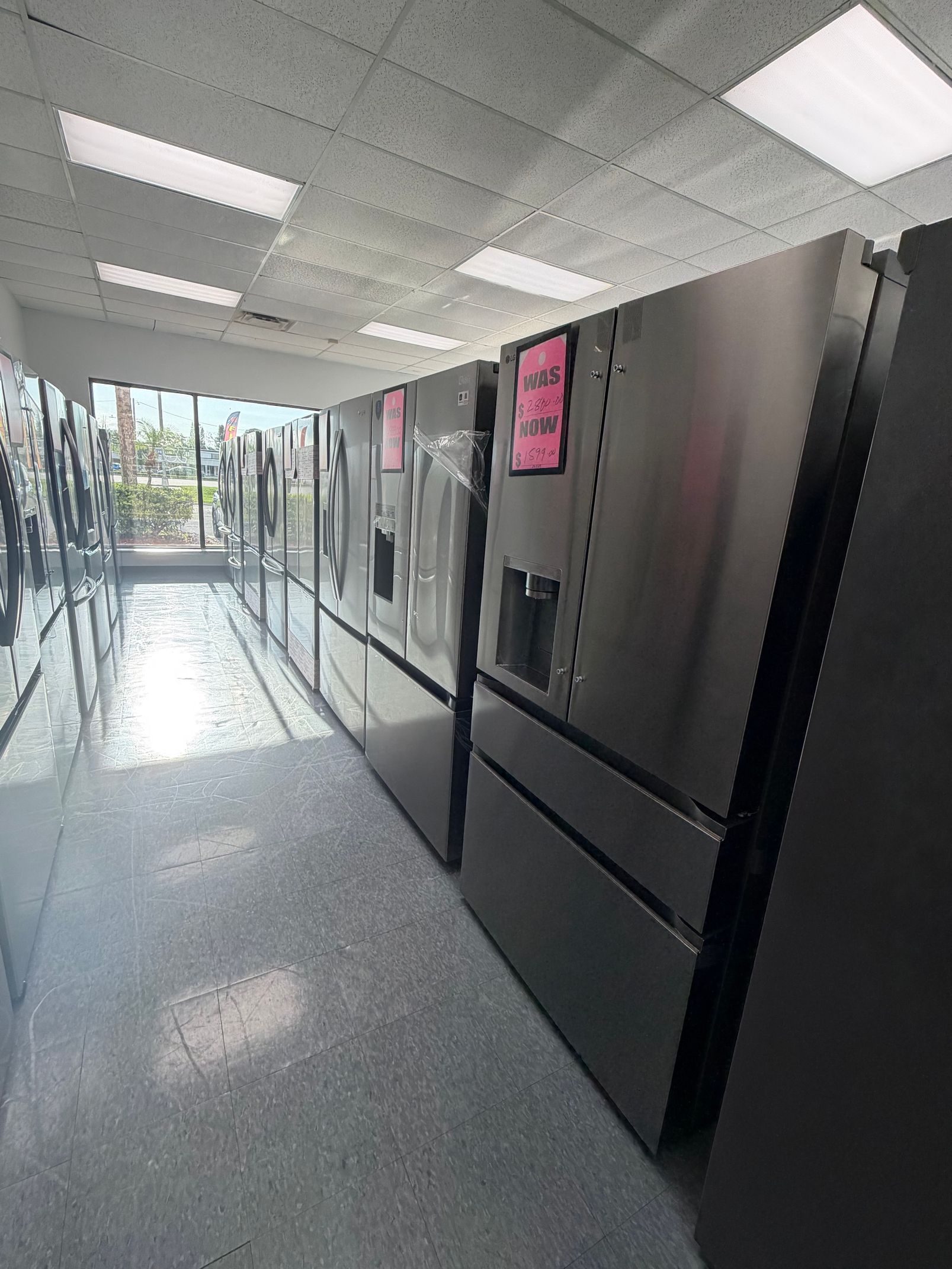 Rows of stainless steel refrigerators in a bright cafeteria hallway near a glass entrance.