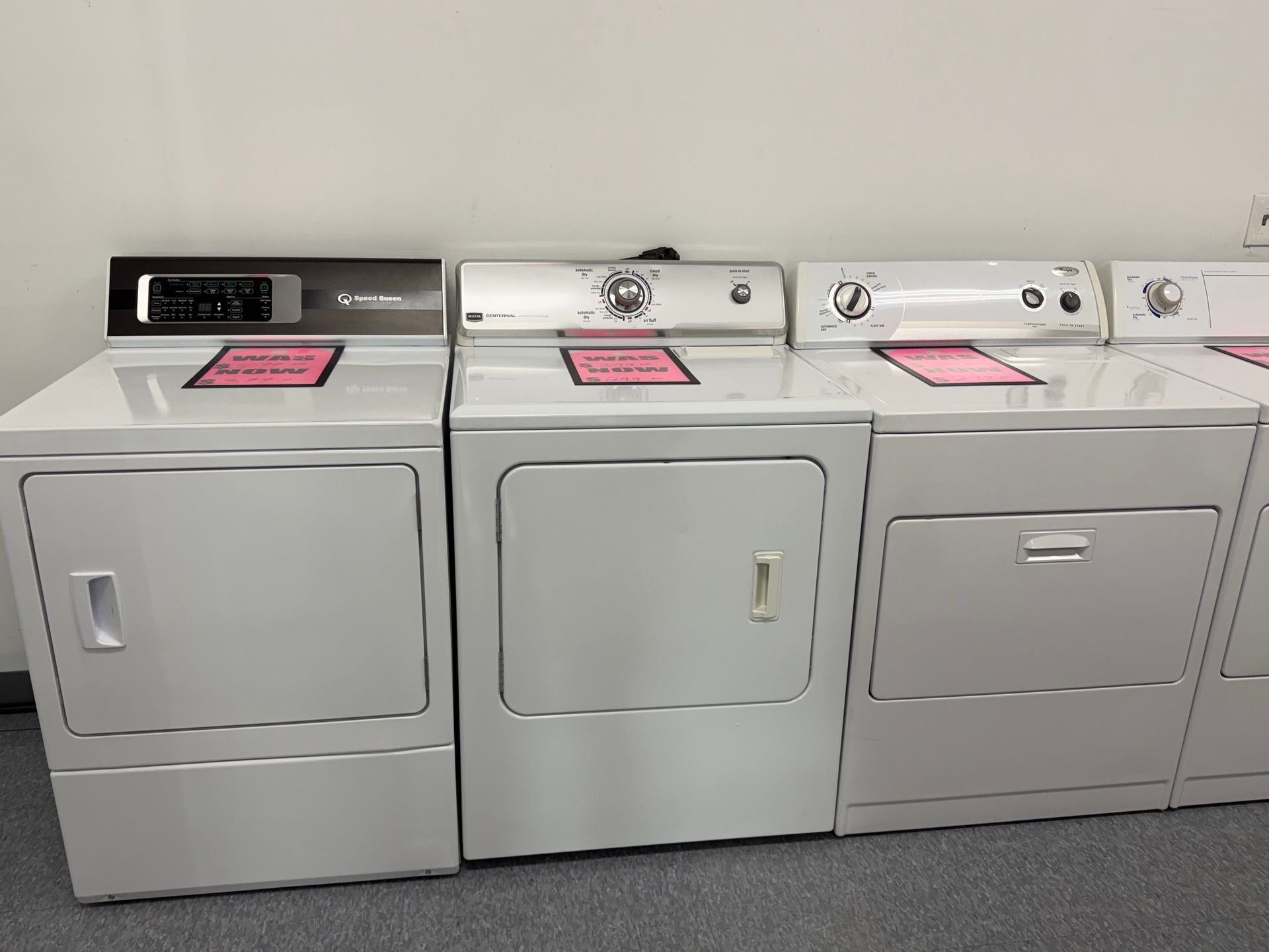 Three side-by-side laundry machines, likely dryers, in a laundromat with pink signs on top.