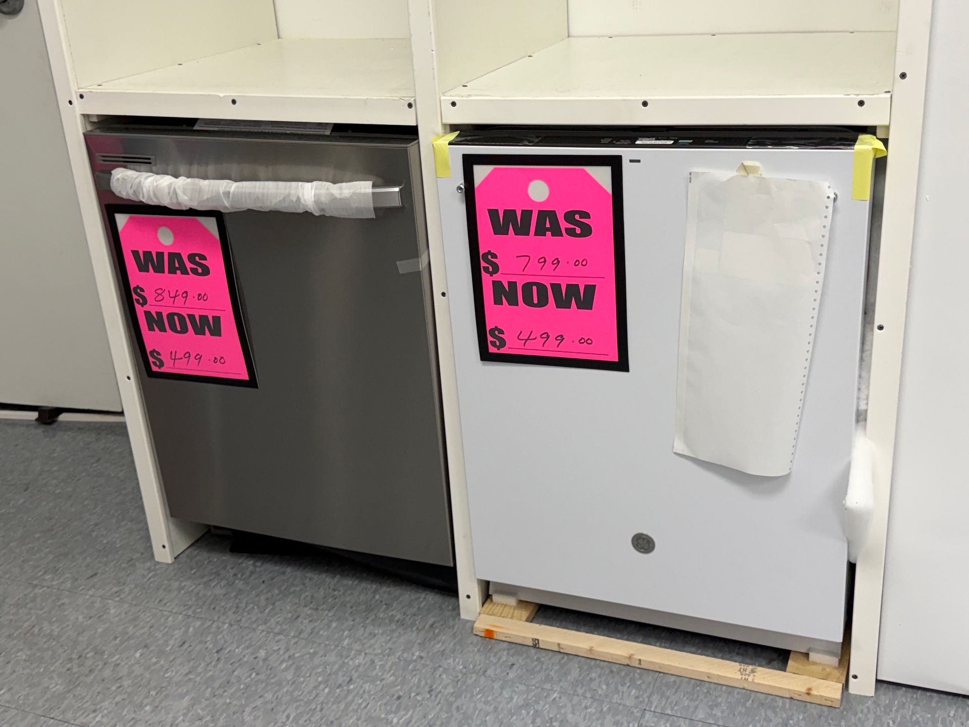 Two dishwashers side by side, one stainless and one white, each with a pink “WAS S NOW” sign.