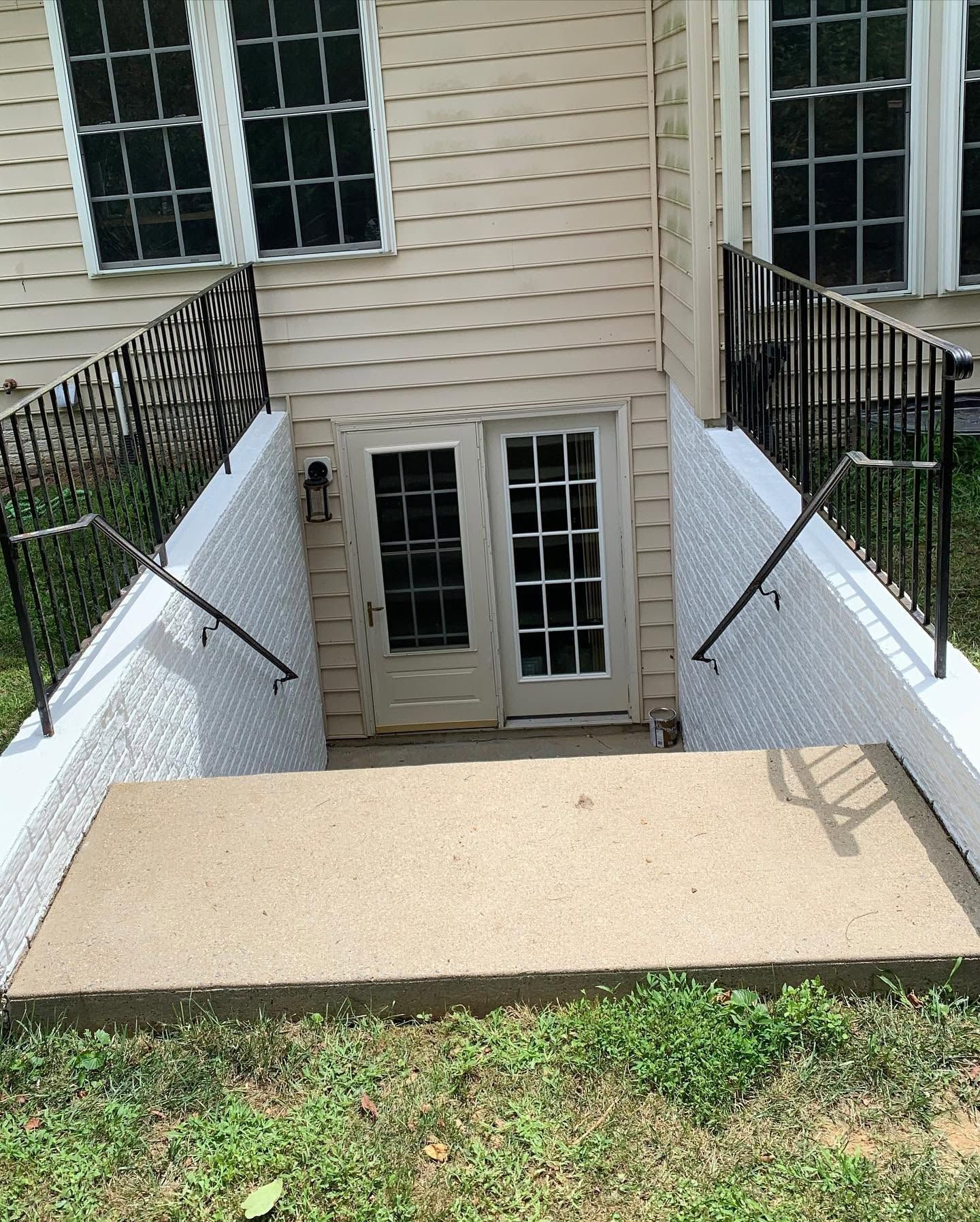 Concrete stair ramp to basement doors with black handrails and white sidewalls beside a beige house