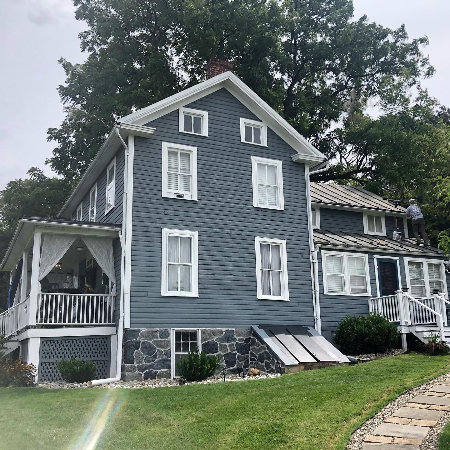 Gray house with white trim and stone foundation on a grassy slope, with two workers on the roof