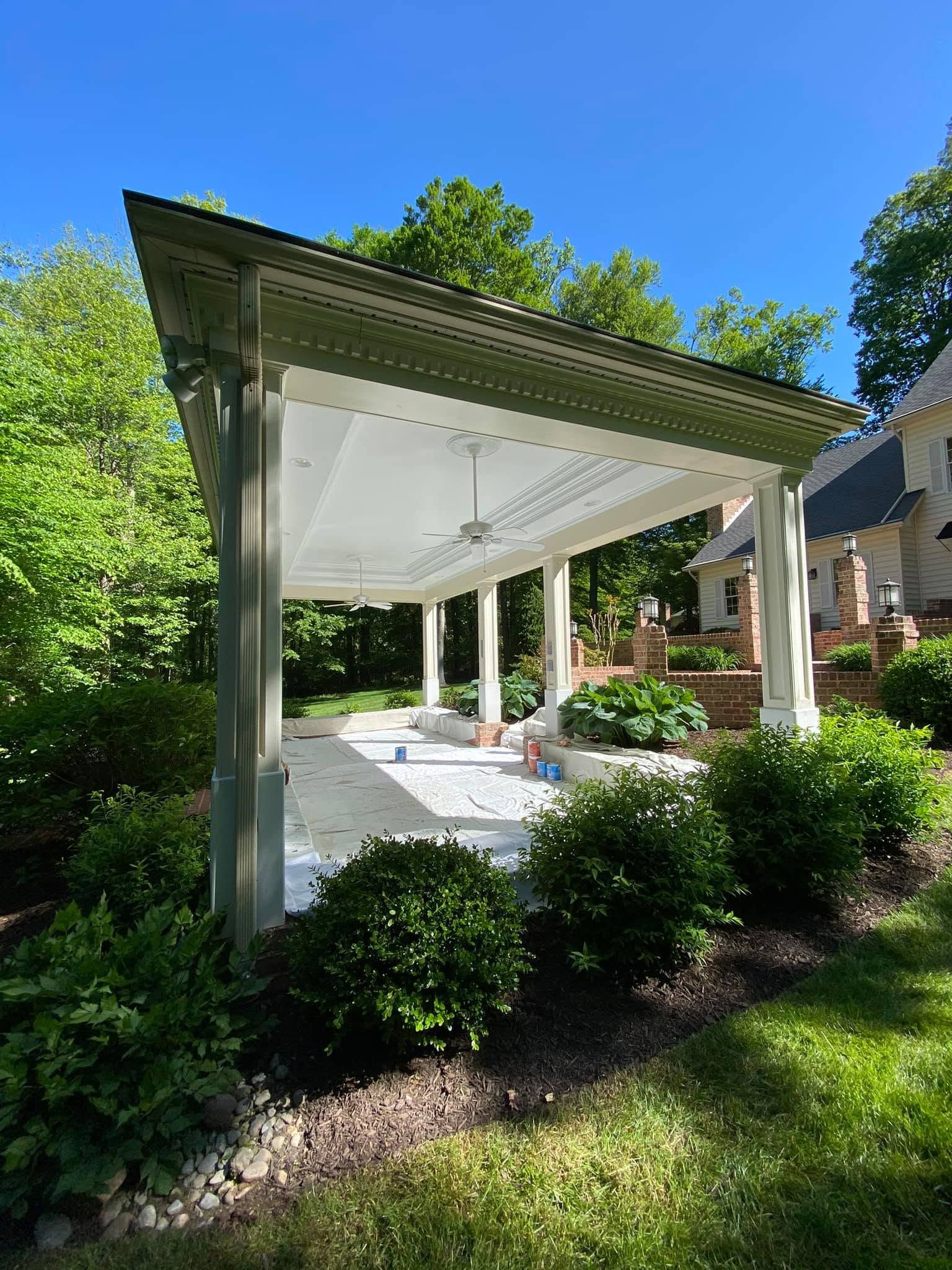 White outdoor pavilion with a sloped roof, surrounded by green shrubs and trees under a blue sky.