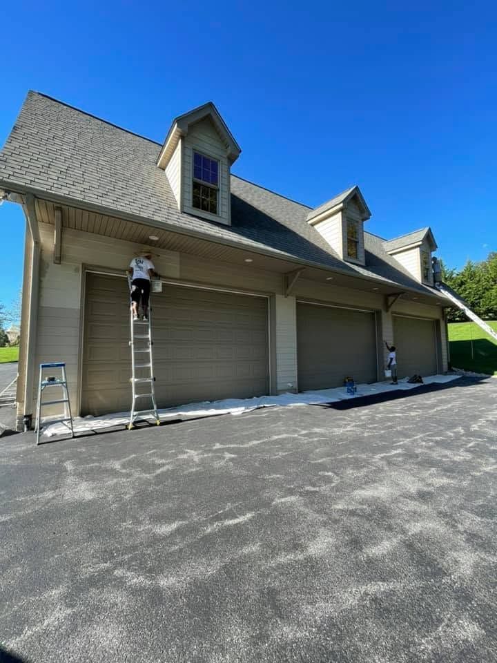 Tan house with three garage doors, two people on ladders, and a wide paved driveway under a blue sky