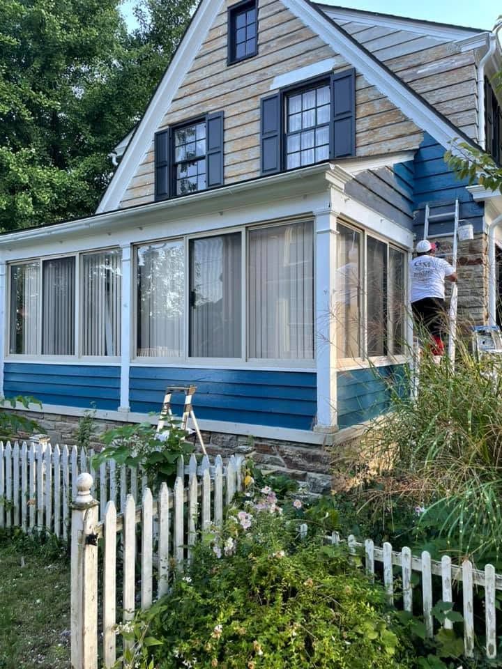 House with blue-and-white siding being painted, with a worker on a ladder by the porch.