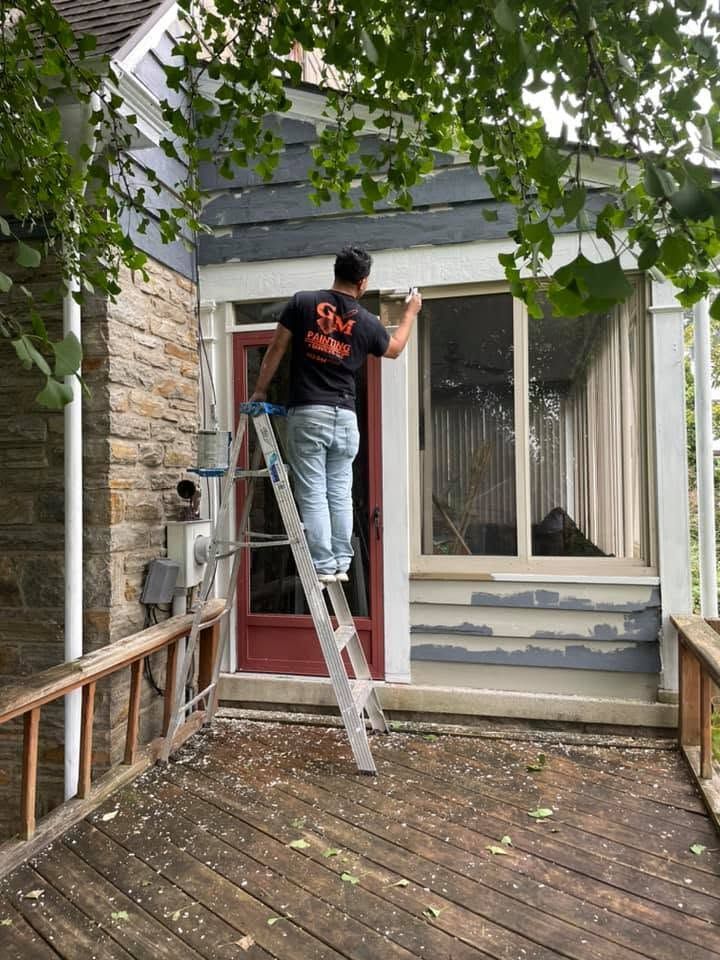 Person on a ladder painting a red front door beside a house with a stone wall and white trim