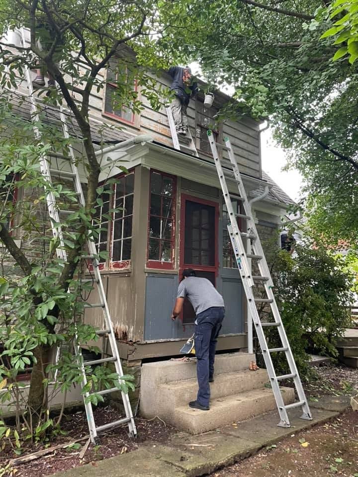 Two workers on ladders repair the roof and front of a small house surrounded by trees.